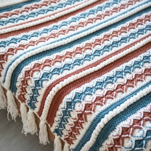 May include: A crocheted throw blanket with a striped pattern in white, teal, and rust colors. The blanket has a fringe border and is draped over a bed.