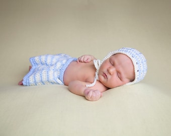 May include: A newborn baby wearing a blue and white striped crocheted outfit and a white crocheted bonnet is sleeping on a light yellow blanket.