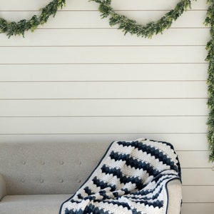 May include: A white and navy blue crocheted throw blanket draped over a light gray couch. The blanket has a chevron pattern and is made with a soft yarn. The couch is in front of a white wood paneled wall with a green garland hanging above.