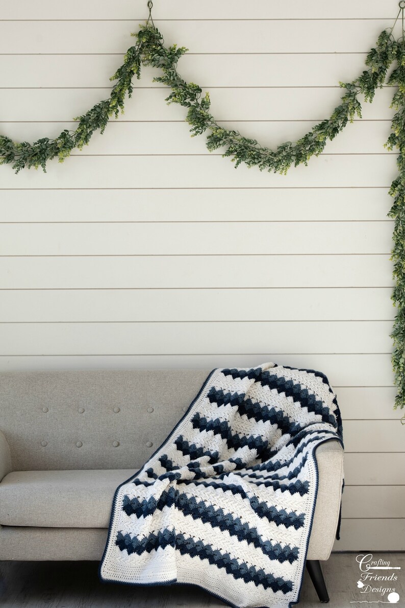 May include: A white and navy blue crocheted throw blanket draped over a light gray couch. The blanket has a chevron pattern and is made with a soft yarn. The couch is in front of a white wood paneled wall with a green garland hanging above.