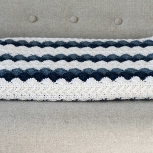 May include: A white and blue crocheted blanket with a chevron pattern. The blanket is folded and sitting on a light gray couch.