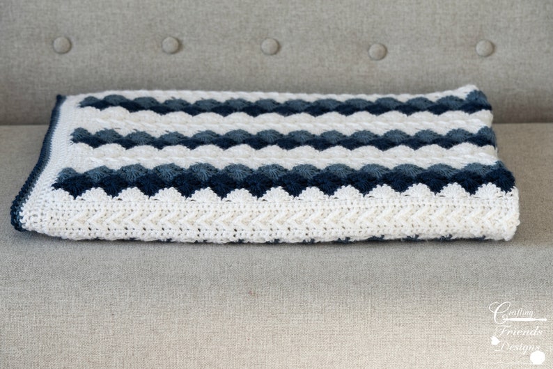 May include: A white and blue crocheted blanket with a chevron pattern. The blanket is folded and sitting on a light gray couch.