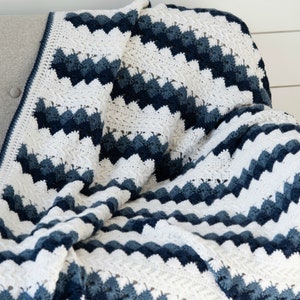 May include: A crocheted blanket with a chevron pattern in shades of blue and white. The blanket is folded and draped over a gray couch.