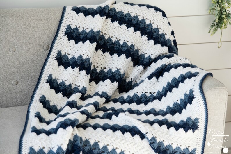 May include: A crocheted blanket with a chevron pattern in shades of blue and white. The blanket is folded and draped over a gray couch.