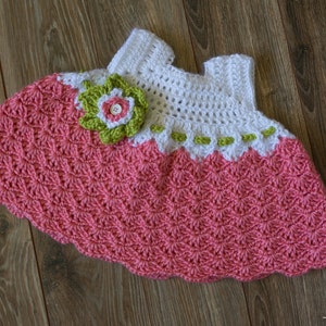Peut inclure: Une robe de b&eacute;b&eacute; en crochet avec un corsage rose et blanc et une jupe rose. La robe est orn&eacute;e d'une fleur verte et blanche.