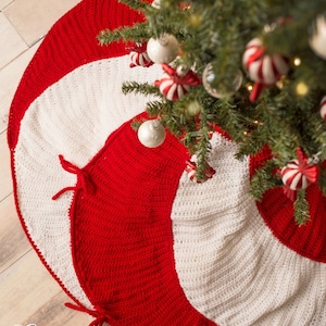 Può includere: Una gonna per albero di Natale in crochet rosso e bianco con un motivo a bastoncino di zucchero. La gonna è realizzata con un motivo a spirale e ha un bordo bianco. La gonna è mostrata su un pavimento di legno con un albero di Natale sullo sfondo.
