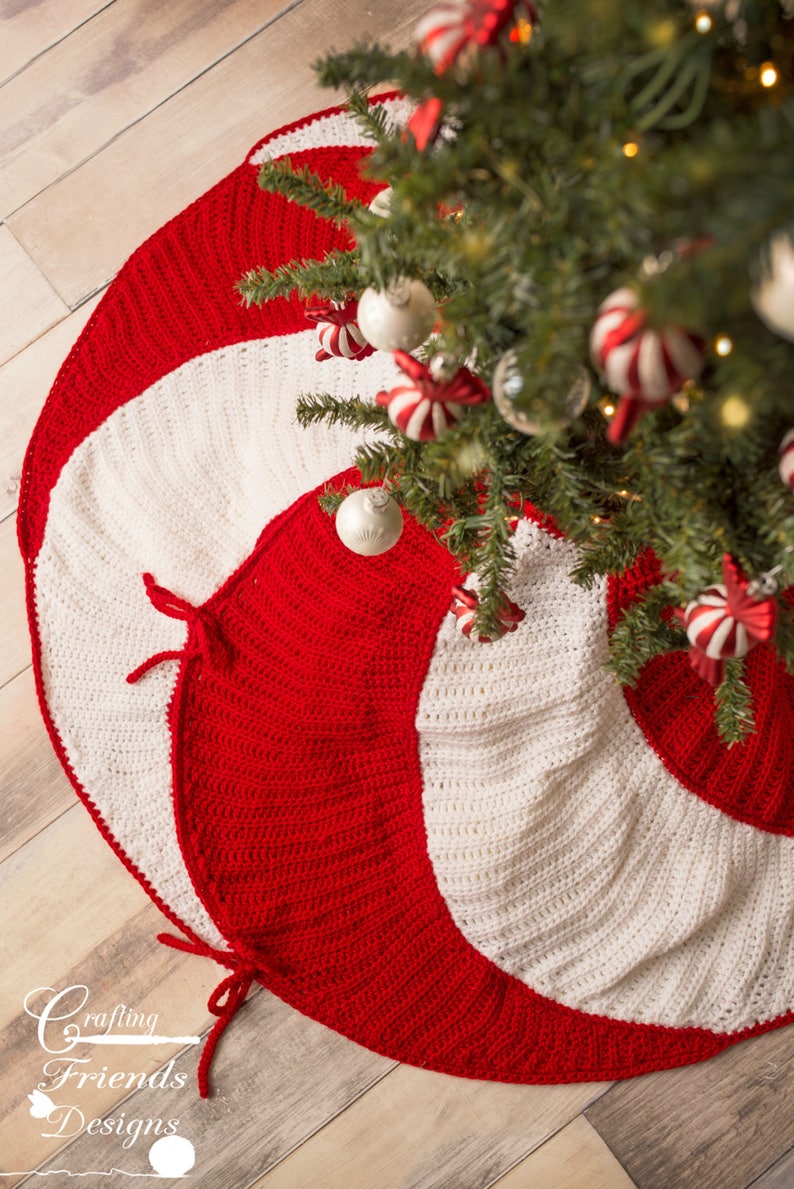 May include: A red and white crocheted Christmas tree skirt with a candy cane design. The skirt is made with a spiral pattern and has a white border. The skirt is shown on a wooden floor with a Christmas tree in the background.