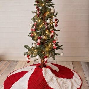 May include: A red and white crocheted Christmas tree skirt with a candy cane design. The skirt is decorated with white and red bows. The skirt is placed under a small Christmas tree decorated with white ornaments and red and white candy cane ornaments.