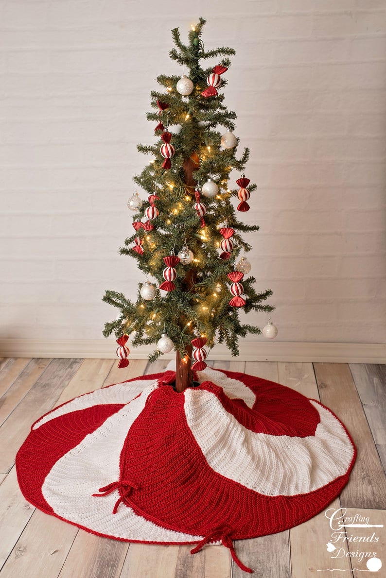 May include: A red and white crocheted Christmas tree skirt with a candy cane design. The skirt is decorated with white and red bows. The skirt is placed under a small Christmas tree decorated with white ornaments and red and white candy cane ornaments.