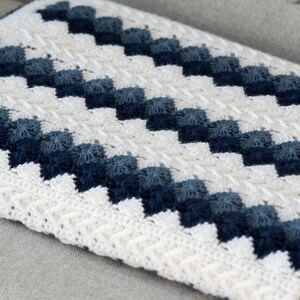 May include: A white and blue crocheted blanket with a chevron pattern. The blanket is made with a variety of shades of blue yarn, creating a gradient effect.
