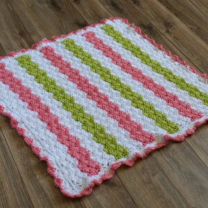 Peut inclure: Une couverture pour b&eacute;b&eacute; en crochet avec un motif chevron en rose, vert et blanc. La couverture a un bord festonn&eacute;.