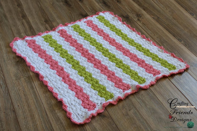 Peut inclure: Une couverture pour b&eacute;b&eacute; en crochet avec un motif chevron en rose, vert et blanc. La couverture a un bord festonn&eacute;.