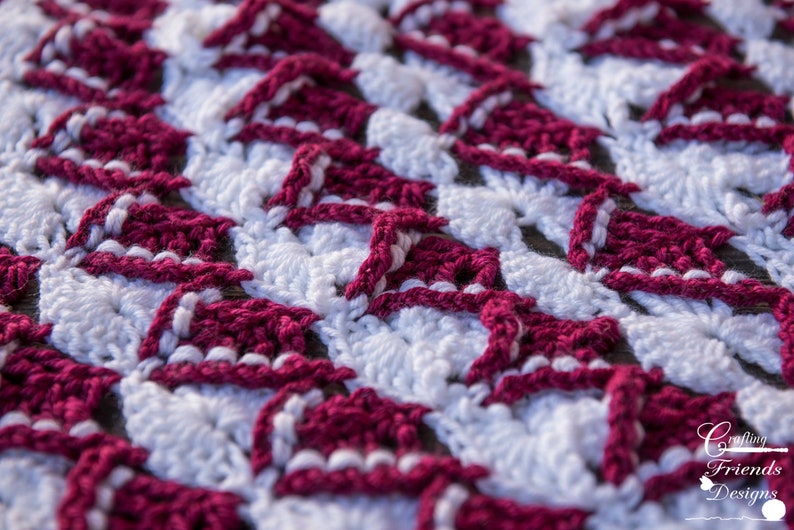 May include: Close-up of a crocheted fabric with a repeating pattern of burgundy and white triangles. The fabric is textured and has a soft, fuzzy appearance.