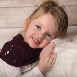 May include: A young girl with blonde hair is wearing a maroon crocheted scarf. She is smiling and looking at the camera. She is sitting on a white fluffy surface.