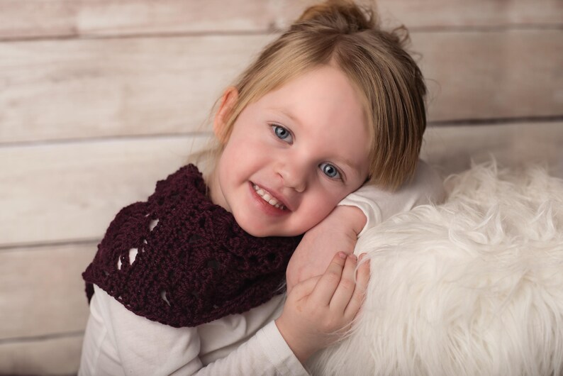 May include: A young girl with blonde hair is wearing a maroon crocheted scarf. She is smiling and looking at the camera. She is sitting on a white fluffy surface.