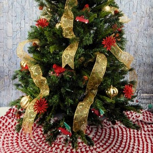 May include: A red and white crocheted Christmas tree skirt with a green Christmas tree decorated with gold ribbon and red ornaments in the background.