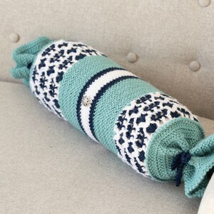 May include: A crocheted pillow in shades of teal, white, and navy blue. The pillow is shaped like a long cylinder and has a decorative knot at one end. The pillow is lying on a light beige couch.