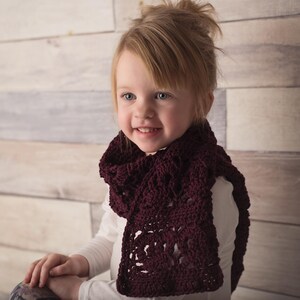 May include: A young girl wearing a burgundy crocheted scarf. The scarf has a lacy pattern and is worn around the neck.