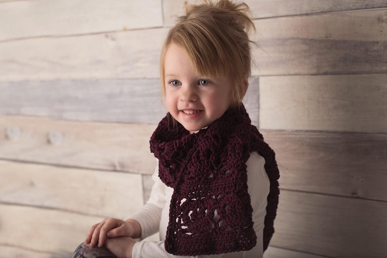 May include: A young girl wearing a burgundy crocheted scarf. The scarf has a lacy pattern and is worn around the neck.