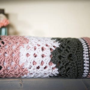 May include: A crocheted blanket with stripes of pink, white, and dark green. The blanket is folded in half and sitting on a wooden surface.