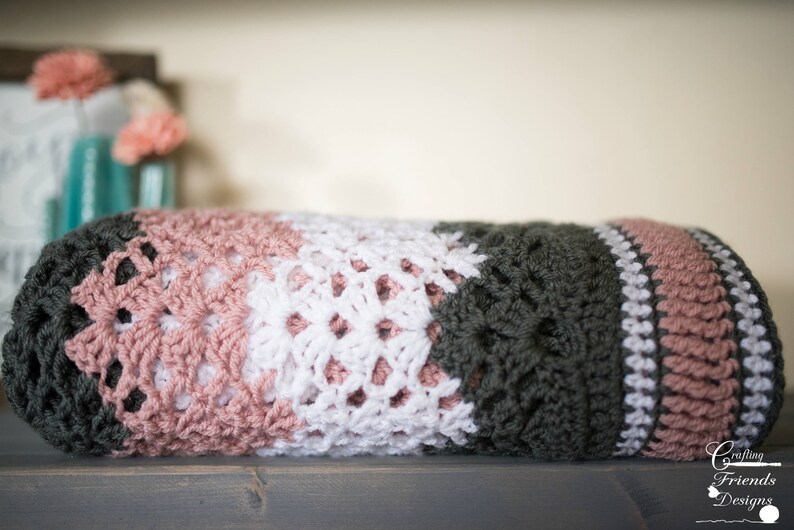 May include: A crocheted blanket with stripes of pink, white, and dark green. The blanket is folded in half and sitting on a wooden surface.