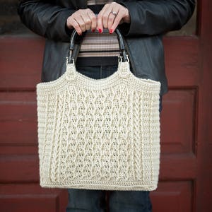 Puede incluir: Un bolso de mano de crochet con un patrón de punto de cable. El bolso es de color blanco roto y tiene asas de cuero marrón.