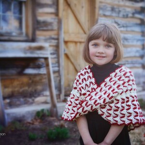 May include: A young girl wearing a red and white crocheted shawl with a black turtleneck shirt. The shawl has a unique pattern and is tied in the front. The girl is standing in front of a wooden building.