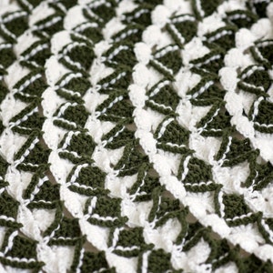 May include: A close-up of a crocheted tree skirt in white and green. The skirt is made with a repeating pattern of triangles and has a scalloped edge.