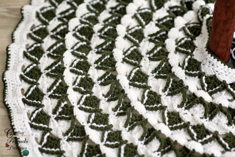 May include: A close-up of a crocheted tree skirt in white and green. The skirt is made with a repeating pattern of triangles and has a scalloped edge.