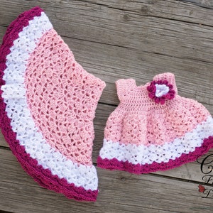 May include: Two crocheted dresses in pink, white, and burgundy. The larger dress has a scalloped hem and the smaller dress has a flower embellishment.