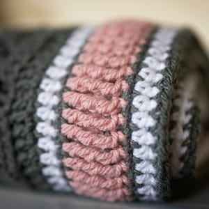 May include: A close-up of a crocheted blanket with stripes of gray, white, and pink yarn. The blanket is rolled up and the edges are visible.