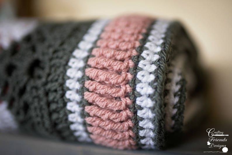 May include: A close-up of a crocheted blanket with stripes of gray, white, and pink yarn. The blanket is rolled up and the edges are visible.