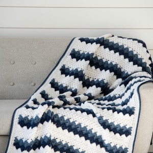 May include: A white and blue crocheted blanket with a chevron pattern is draped over a light gray couch. The blanket has a white border and is made with a soft yarn.