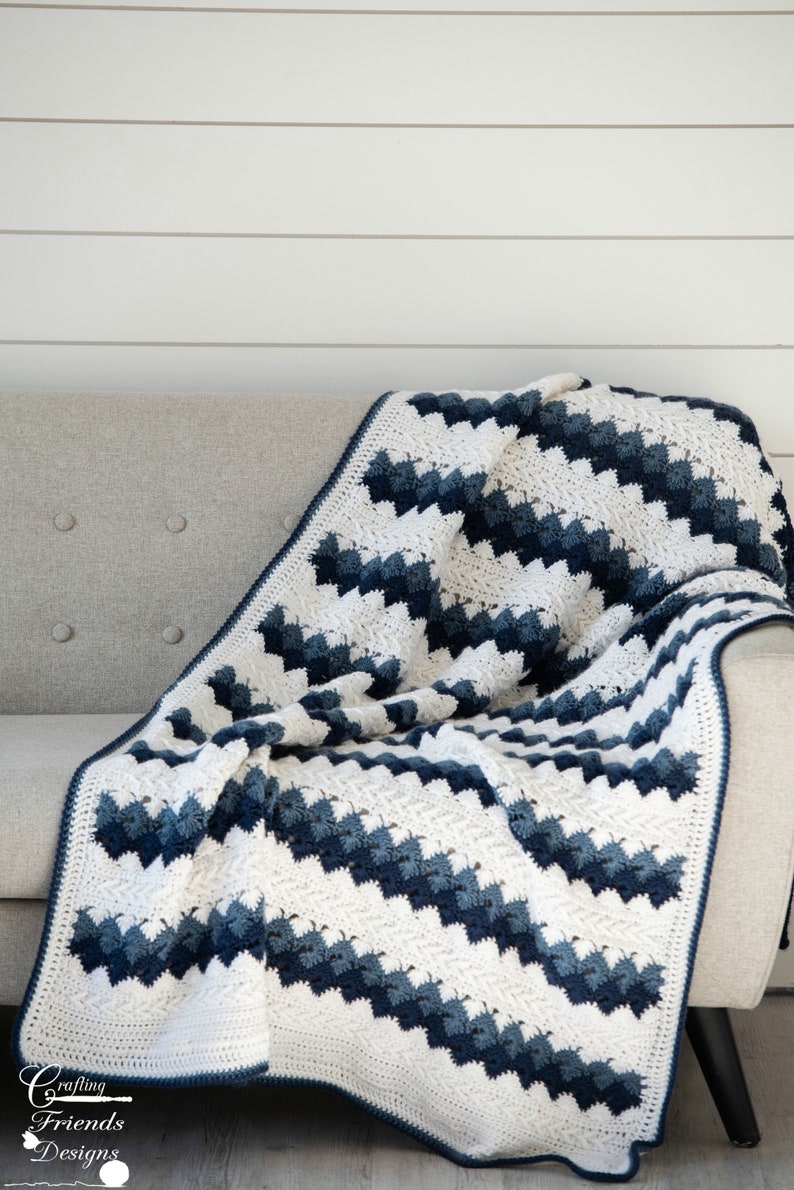 May include: A white and blue crocheted blanket with a chevron pattern is draped over a light gray couch. The blanket has a white border and is made with a soft yarn.