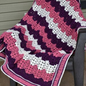 May include: A crocheted blanket with a chevron pattern in shades of pink, purple, and white. The blanket has a border in pink and white.