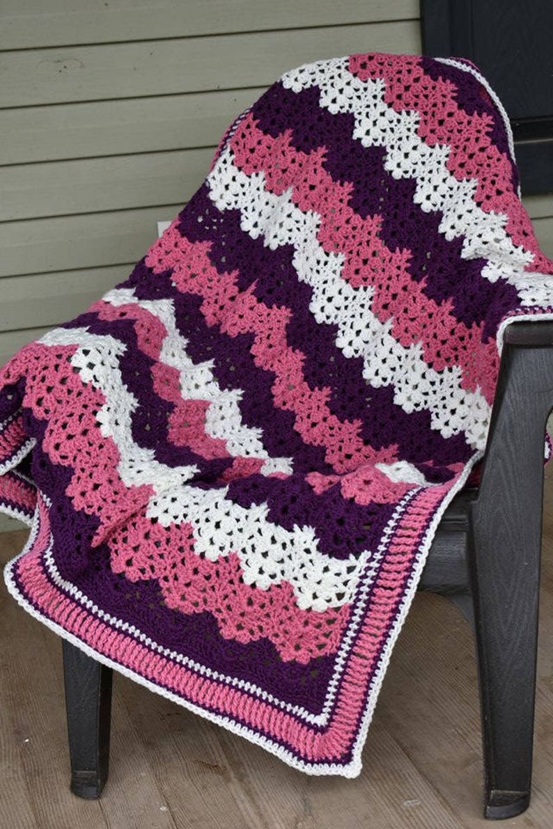 May include: A crocheted blanket with a chevron pattern in shades of pink, purple, and white. The blanket has a border in pink and white.