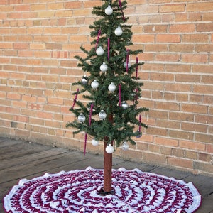 May include: A white and burgundy crochet Christmas tree skirt with a scalloped edge. A small artificial Christmas tree with silver ornaments and a silver star on top is sitting on the skirt.