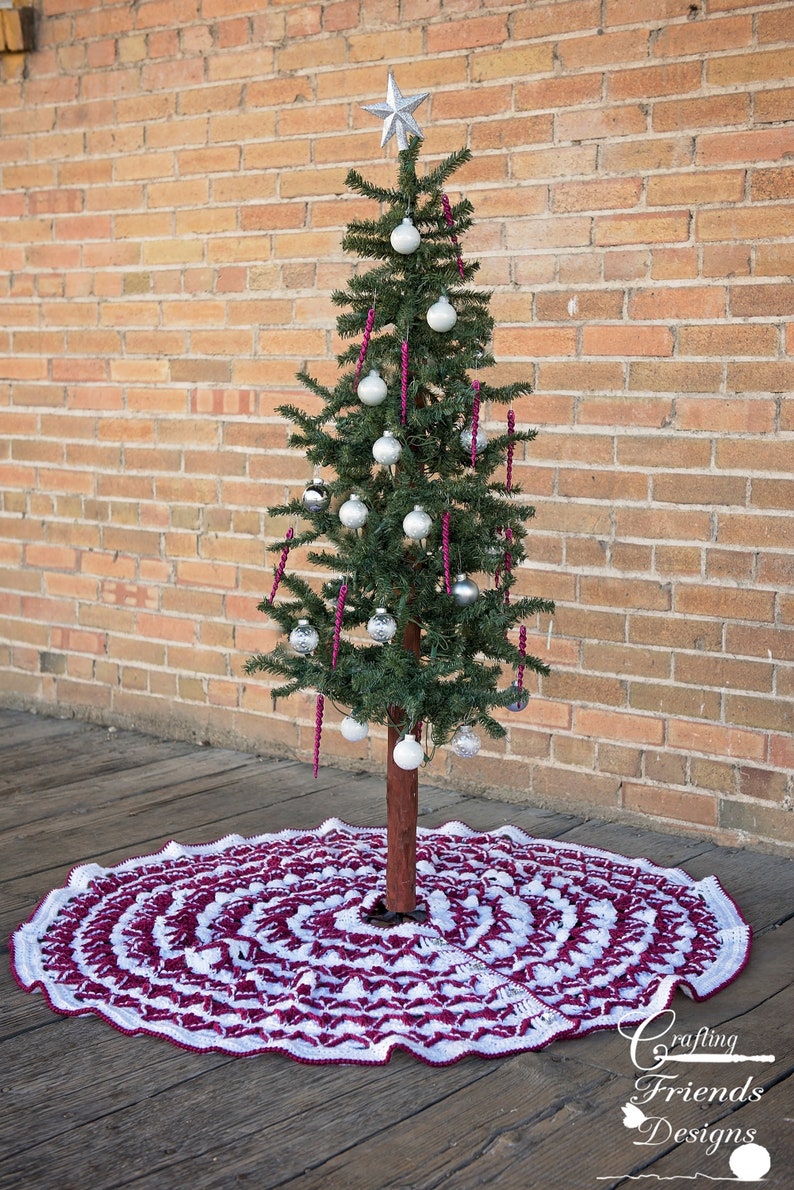 May include: A white and burgundy crochet Christmas tree skirt with a scalloped edge. A small artificial Christmas tree with silver ornaments and a silver star on top is sitting on the skirt.