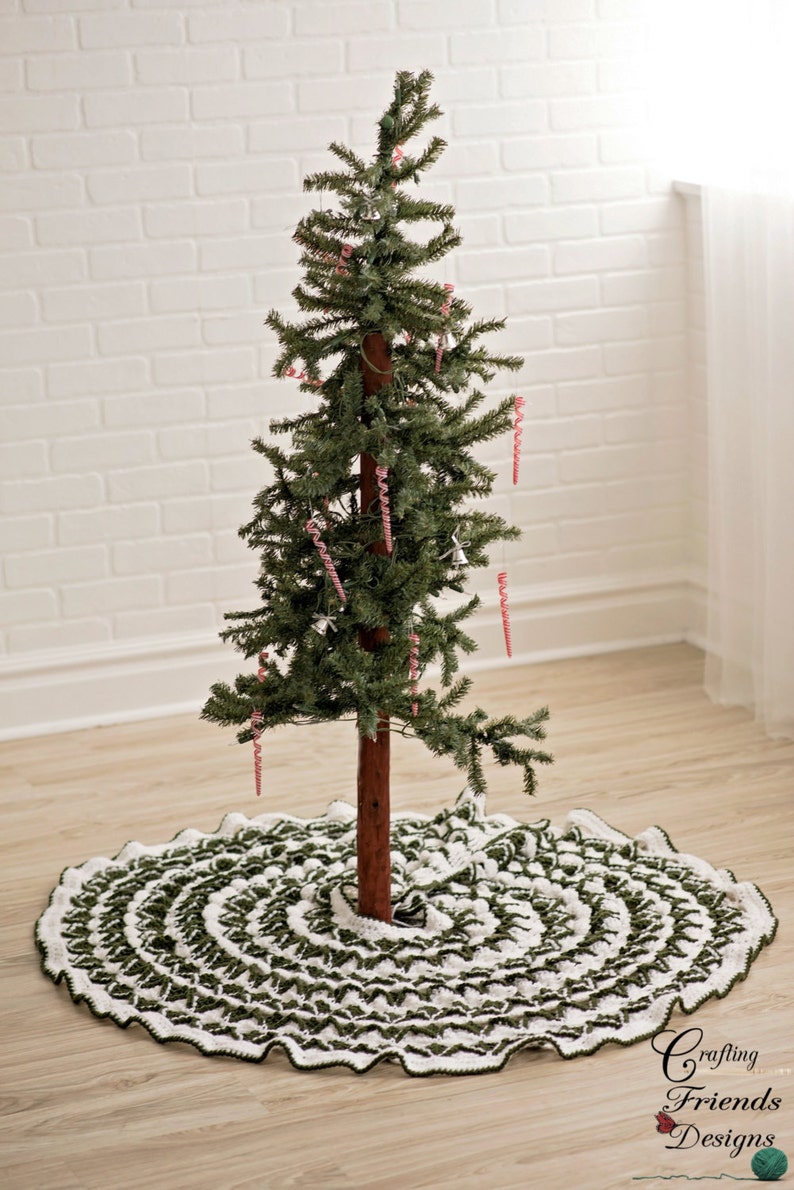 May include: A green and white crocheted Christmas tree skirt with a ruffled edge. The skirt is placed on a wood floor under a small artificial Christmas tree decorated with candy cane ornaments.