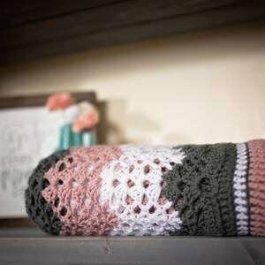 May include: A crocheted blanket with stripes of pink, white, and dark green. The blanket is folded and sitting on a wooden surface.