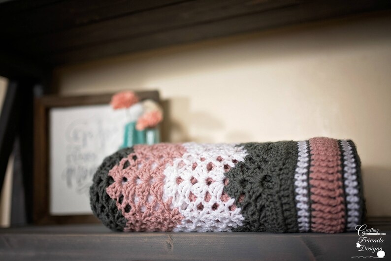 May include: A crocheted blanket with stripes of pink, white, and dark green. The blanket is folded and sitting on a wooden surface.