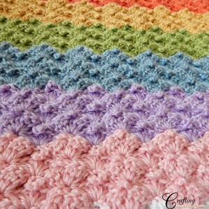 May include: A rainbow crochet blanket with a textured, scalloped stitch pattern. The blanket is made in shades of pink, purple, blue, green, yellow, and orange. The blanket is finished with a white border.