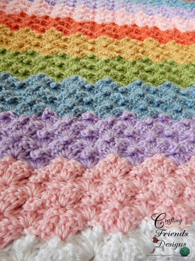 May include: A rainbow crochet blanket with a textured, scalloped stitch pattern. The blanket is made in shades of pink, purple, blue, green, yellow, and orange. The blanket is finished with a white border.