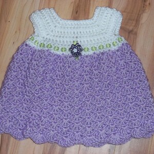 Peut inclure: Une robe de b&eacute;b&eacute; en crochet avec un corsage blanc et une jupe violette. La robe est orn&eacute;e d'une bordure verte et blanche et d'une fleur violette.