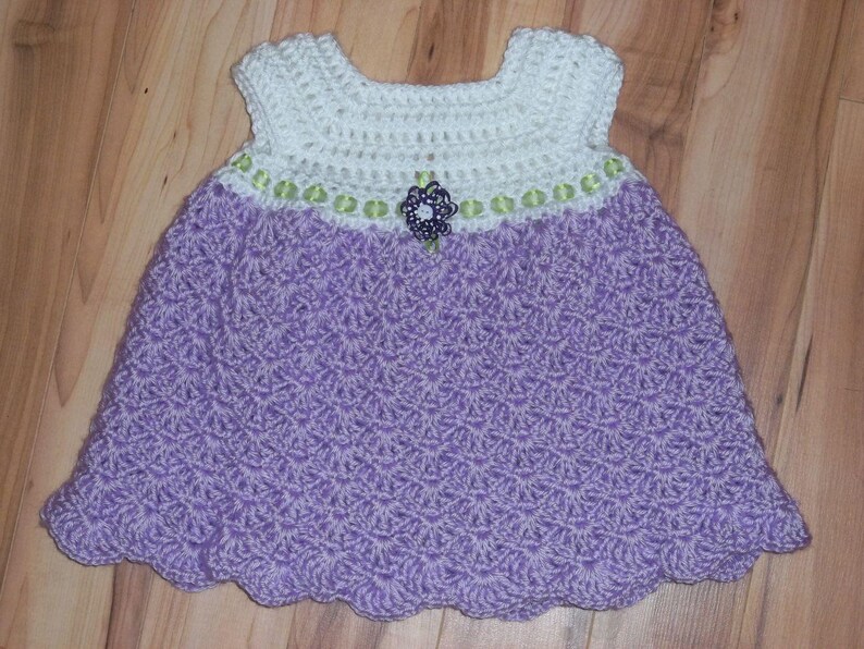 Peut inclure: Une robe de b&eacute;b&eacute; en crochet avec un corsage blanc et une jupe violette. La robe est orn&eacute;e d'une bordure verte et blanche et d'une fleur violette.
