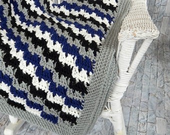 Peut inclure: Une chaise berçante en osier blanc avec une couverture au crochet dans des tons de gris, noir, blanc et bleu. La couverture est pliée sur le dossier de la chaise et drapé sur le siège.