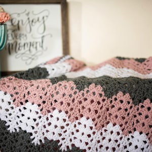 May include: A crocheted blanket with a chevron pattern in shades of pink, white, and gray. The blanket is made with a delicate stitch and has a soft, cozy texture.