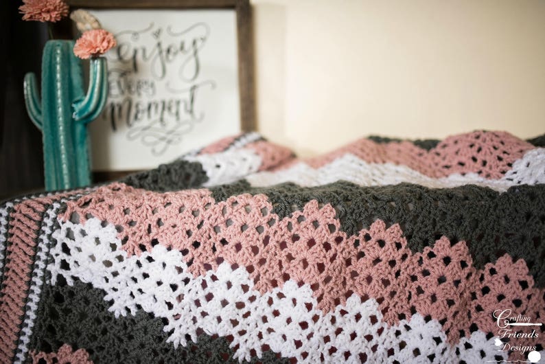 May include: A crocheted blanket with a chevron pattern in shades of pink, white, and gray. The blanket is made with a delicate stitch and has a soft, cozy texture.