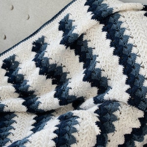 May include: A white and blue crocheted blanket with a chevron pattern. The blanket is made with a soft, textured yarn and has a fringed edge.