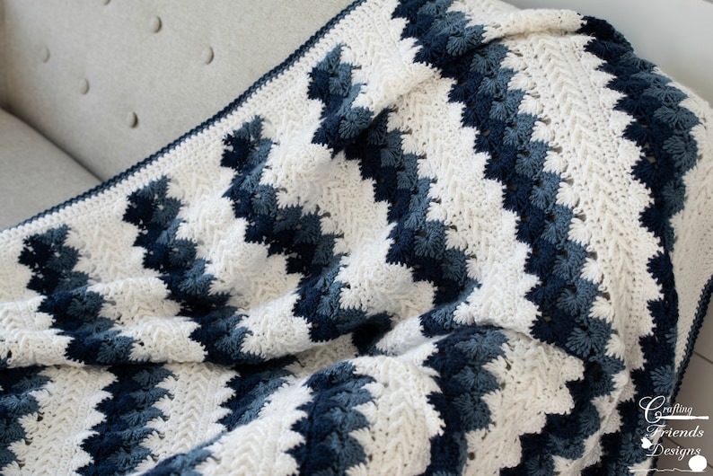 May include: A white and blue crocheted blanket with a chevron pattern. The blanket is made with a soft, textured yarn and has a fringed edge.